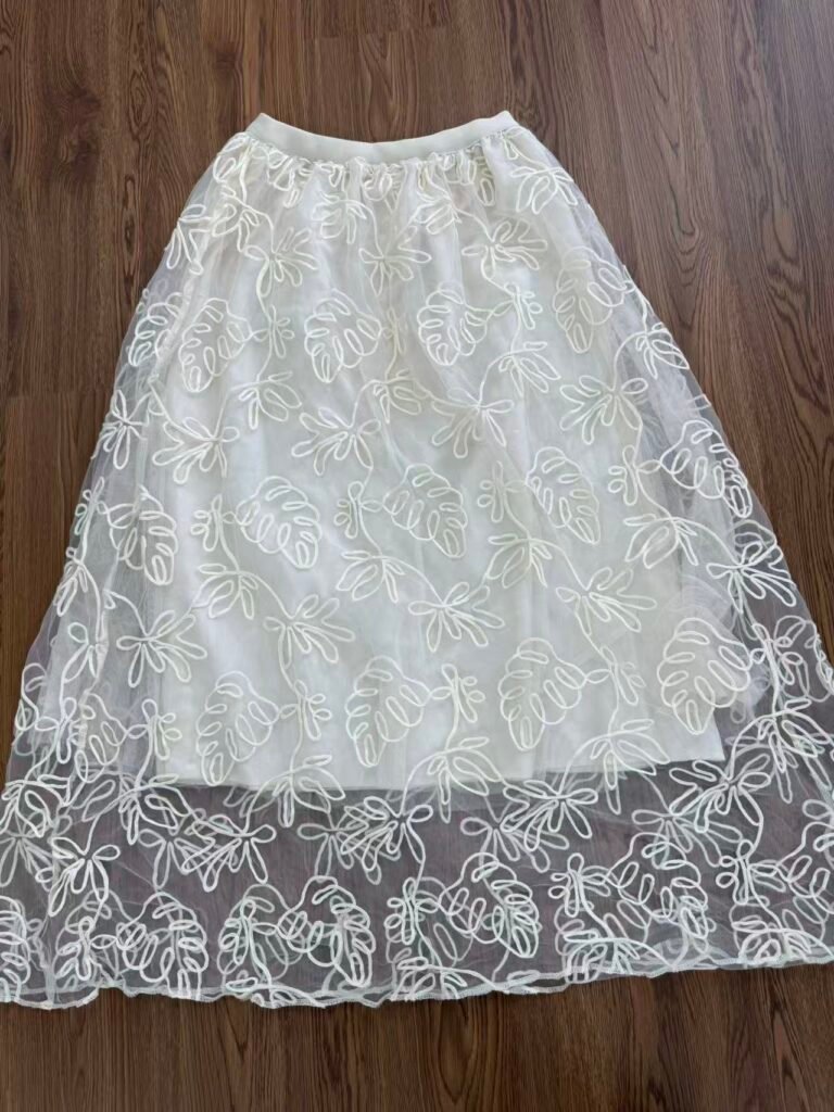A flat lay of a white mesh maxi skirt featuring an intricate leaf-pattern embroidery, showcasing a lightweight and breezy resort-wear aesthetic.