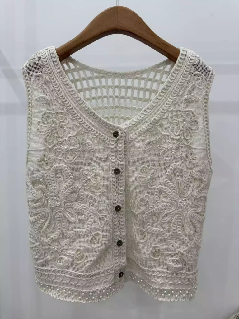 Close-up of a cream-colored sleeveless top with V-neck, featuring intricate floral and swirl embroidery, pearl and bead embellishments, and wooden buttons, hanging on a wooden hanger.