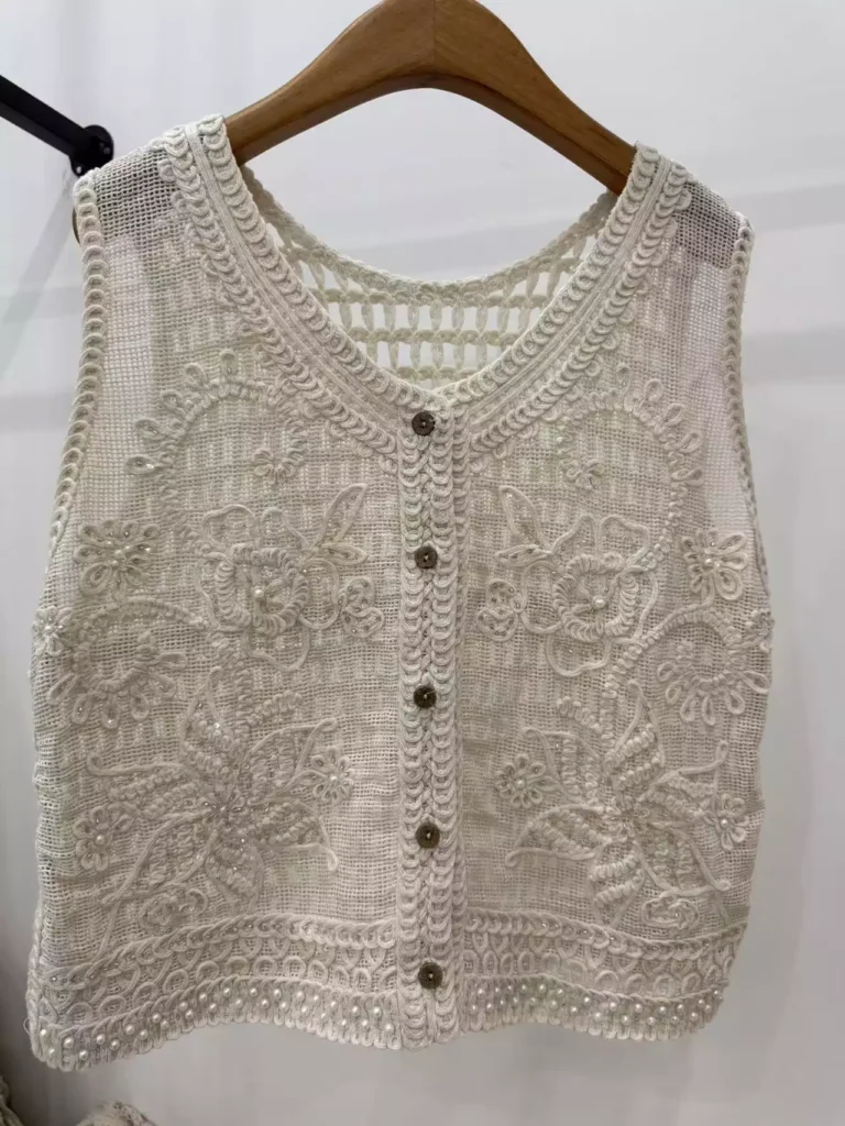 A cream-colored sleeveless vest with intricate 3D floral embroidery, bead accents, a V-neck, button-front, and faux pearl-trimmed hem, displayed on a wooden hanger against a white background.