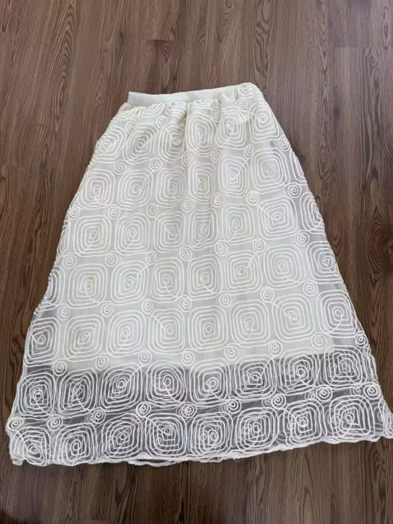 Cream-colored midi skirt with intricate spiral square embroidery, laid flat on a warm wooden floor, showcasing its unique pattern and sheer texture.