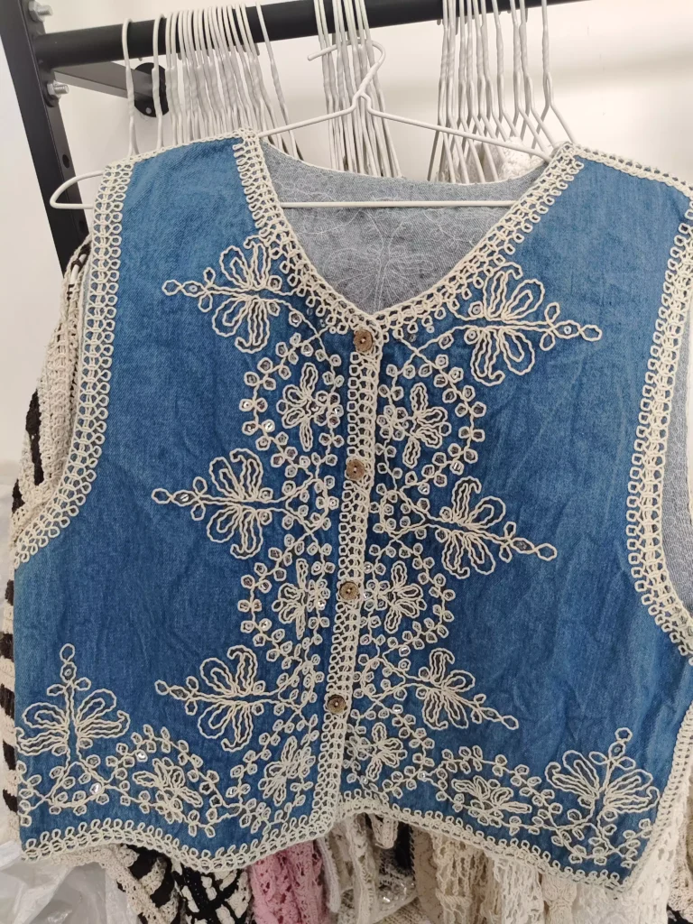 A hanger shot of a blue denim floral embroidery vest, showcasing intricate white embroidery textures, sequin accents, and wooden button details.