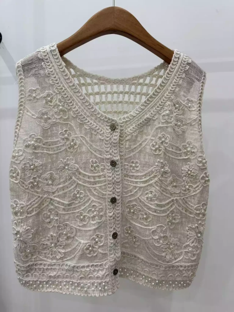 A hanger shot of a white crochet vest, showcasing intricate swirl-pattern embroidery, pearl embellishments, and a button-front design.