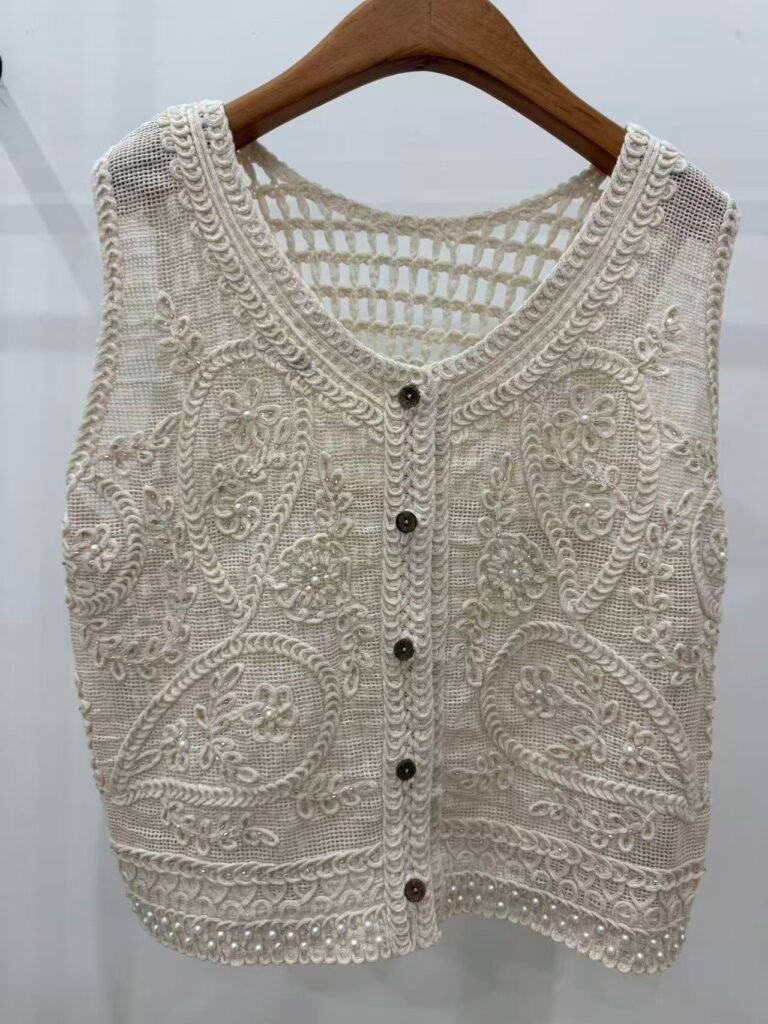 A hanger shot of an ivory floral embroidery crochet vest, showcasing fine mesh base, paisley embroidery textures, pearl embellishments, and button details.