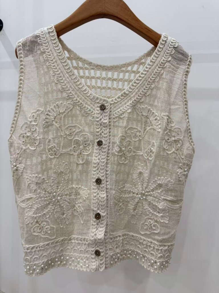 A hanger shot of a white crochet vest, showcasing exquisite floral embroidery, pearl accents, and a button-front design.