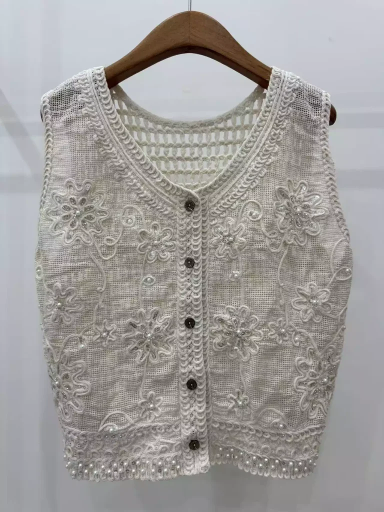 A hanger shot of a white crochet vest, showcasing exquisite floral embroidery, pearl accents, and a button-front design.