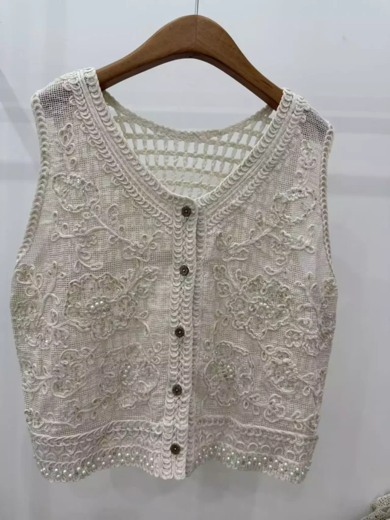 A close-up hanger shot of a white crochet vest, showcasing intricate floral embroidery textures, pearl embellishments, and button-front design.