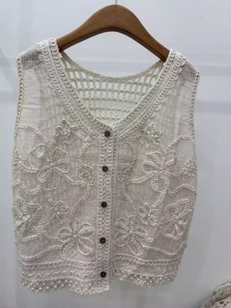 A hanger shot of a white floral embroidery crochet vest, showcasing intricate embroidery textures, pearl embellishments, and button-front design.