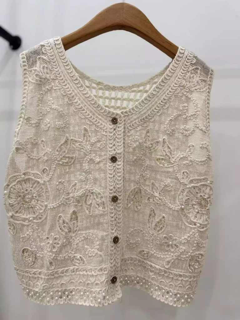A hanger shot of a white crochet vest, showcasing intricate floral embroidery, pearl embellishments, and vintage-style buttons.