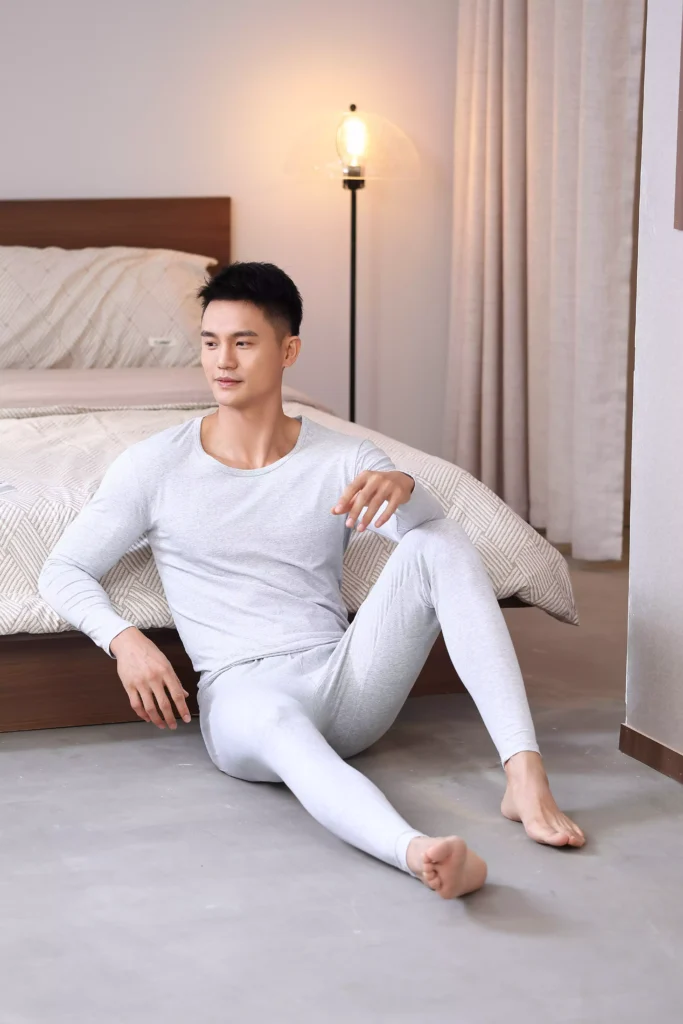 A man wearing a grey thermal underwear set, relaxing on the floor in a bedroom, highlighting the comfortable fit and casual style of the loungewear.