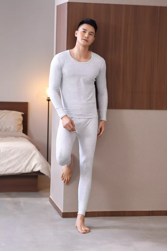 A man wearing a grey thermal underwear set, including a long-sleeved top and matching leggings, posing in a modern bedroom setting.