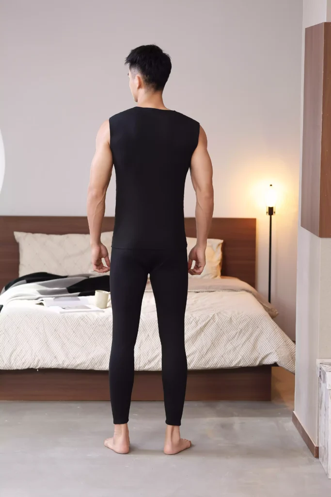 Back view of a man wearing a black sleeveless thermal undershirt and matching thermal leggings, showcasing a sleek, athletic fit in a bedroom environment.