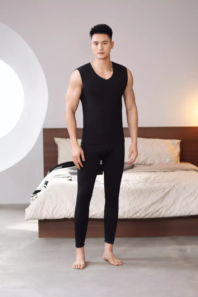 Front view of a man modeling a black sleeveless thermal undershirt and matching leggings in a modern bedroom setting.