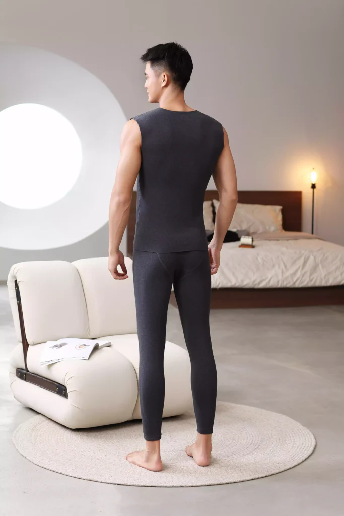 Back view of a man modeling a dark grey sleeveless thermal set in a contemporary bedroom setting, highlighting the slim and comfortable fit.