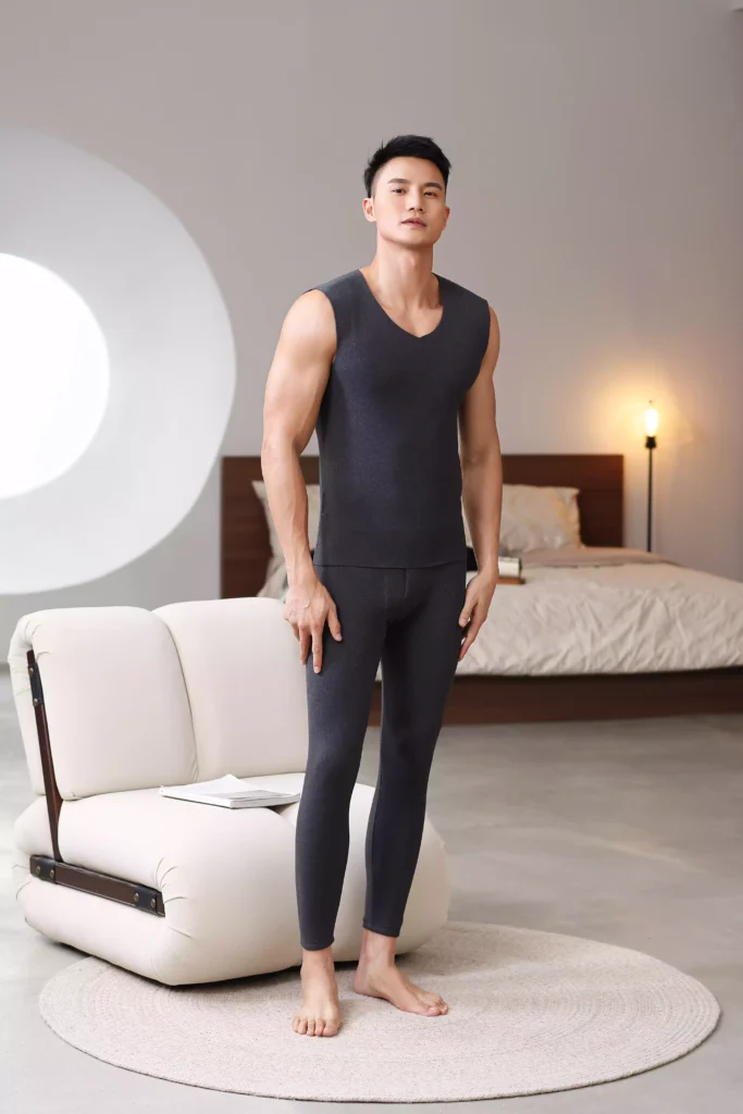 Full-body front view of a man wearing a charcoal dark grey sleeveless thermal undershirt and matching leggings in a stylish, modern bedroom.