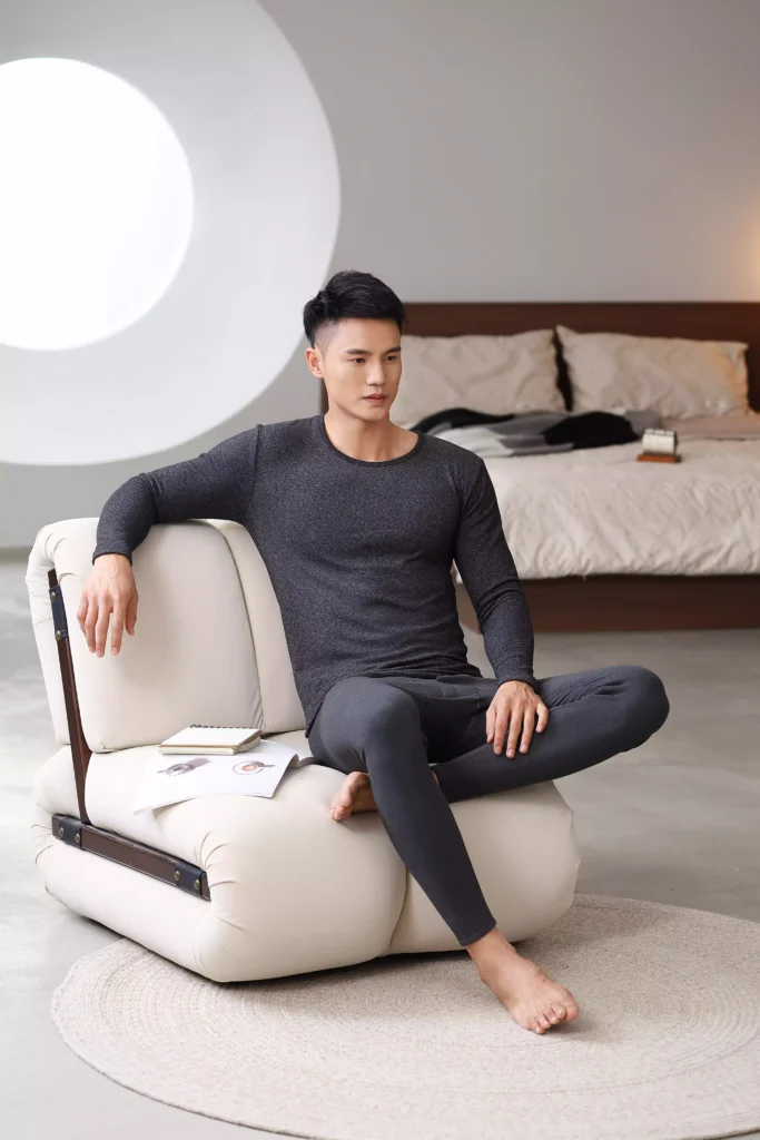 A man sitting and relaxing while wearing a dark grey thermal underwear set, demonstrating comfort and fabric flexibility.