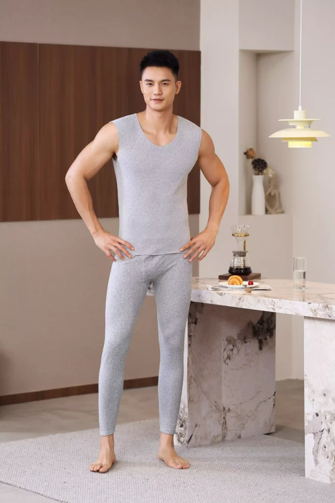 Full-body front view of a man modeling a light grey sleeveless thermal undershirt and matching leggings in a modern home interior.