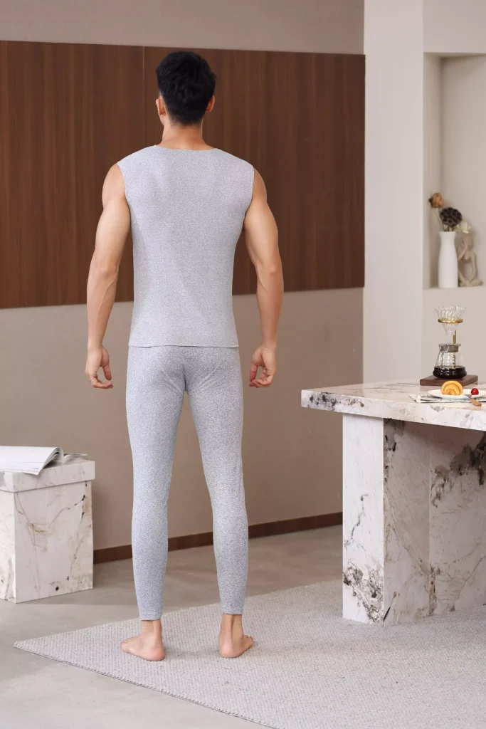 Back view of a man wearing a light grey sleeveless thermal undershirt and matching thermal leggings, showcasing the snug and comfortable fit in a minimalist interior.
