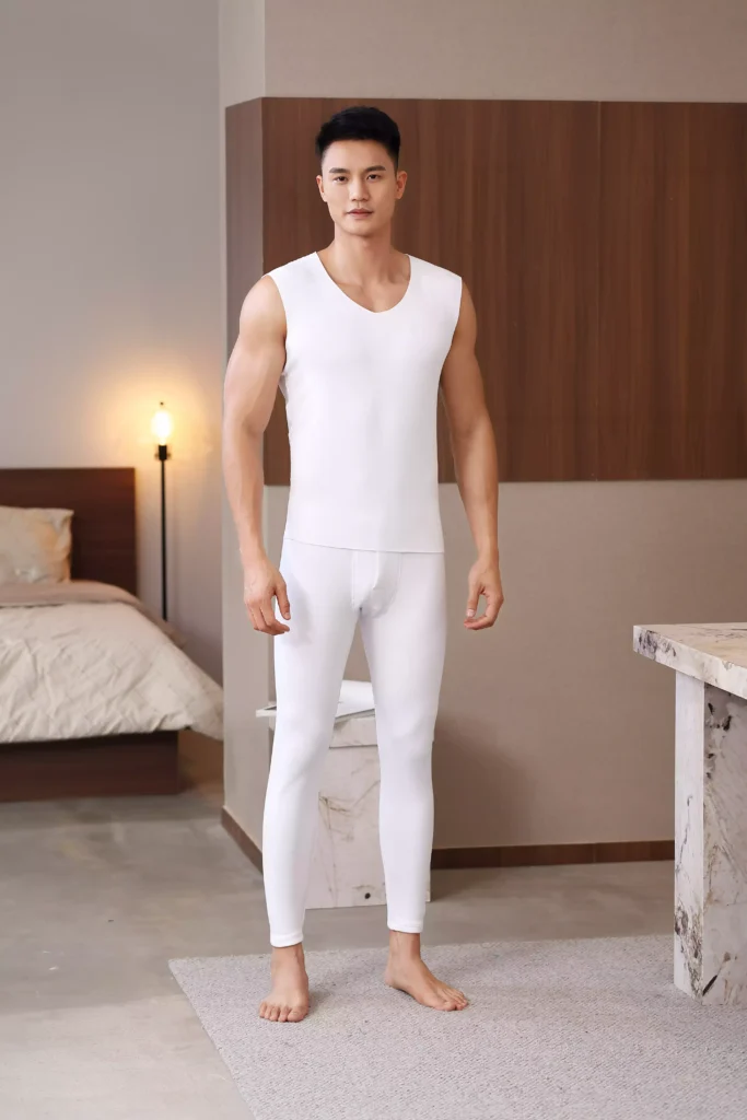 Front view of a man wearing a white sleeveless thermal undershirt and matching leggings, standing in a comfortable bedroom setting.