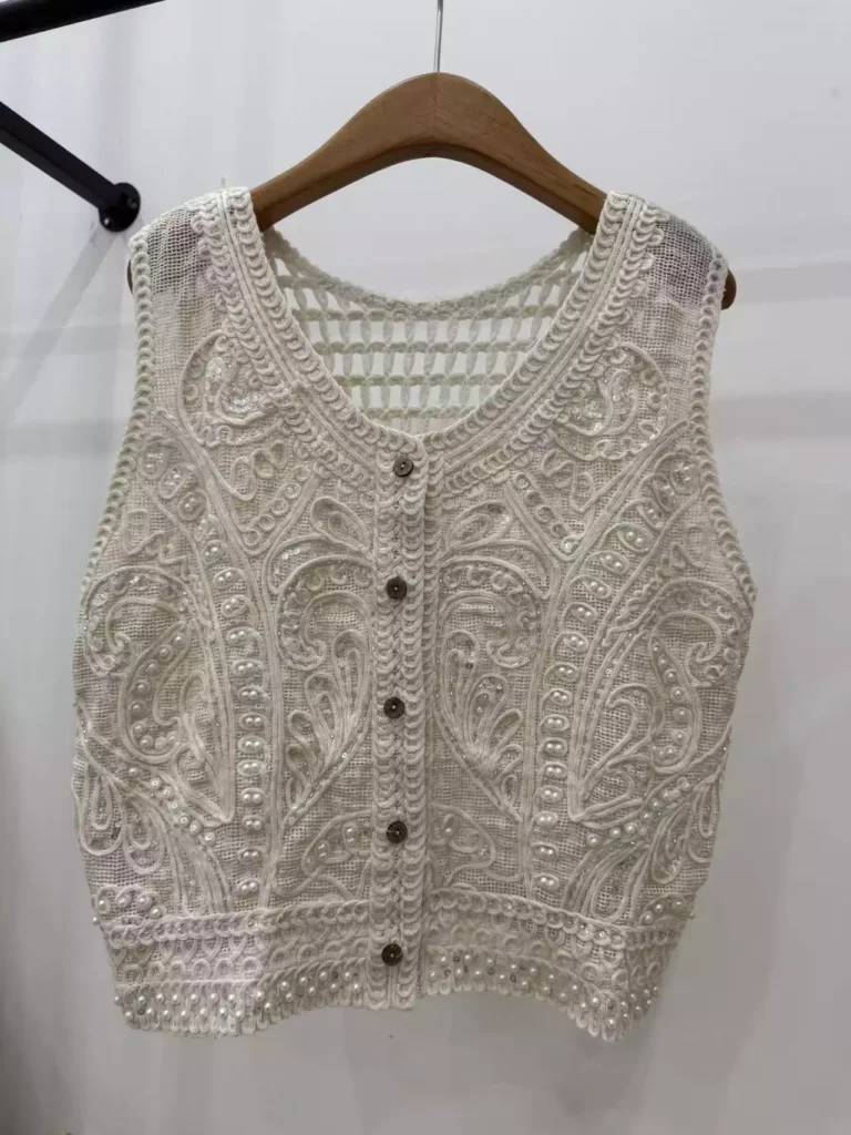 A hanger shot of a white paisley-pattern crochet vest, showcasing intricate pearl and sequin details with a button-front design.