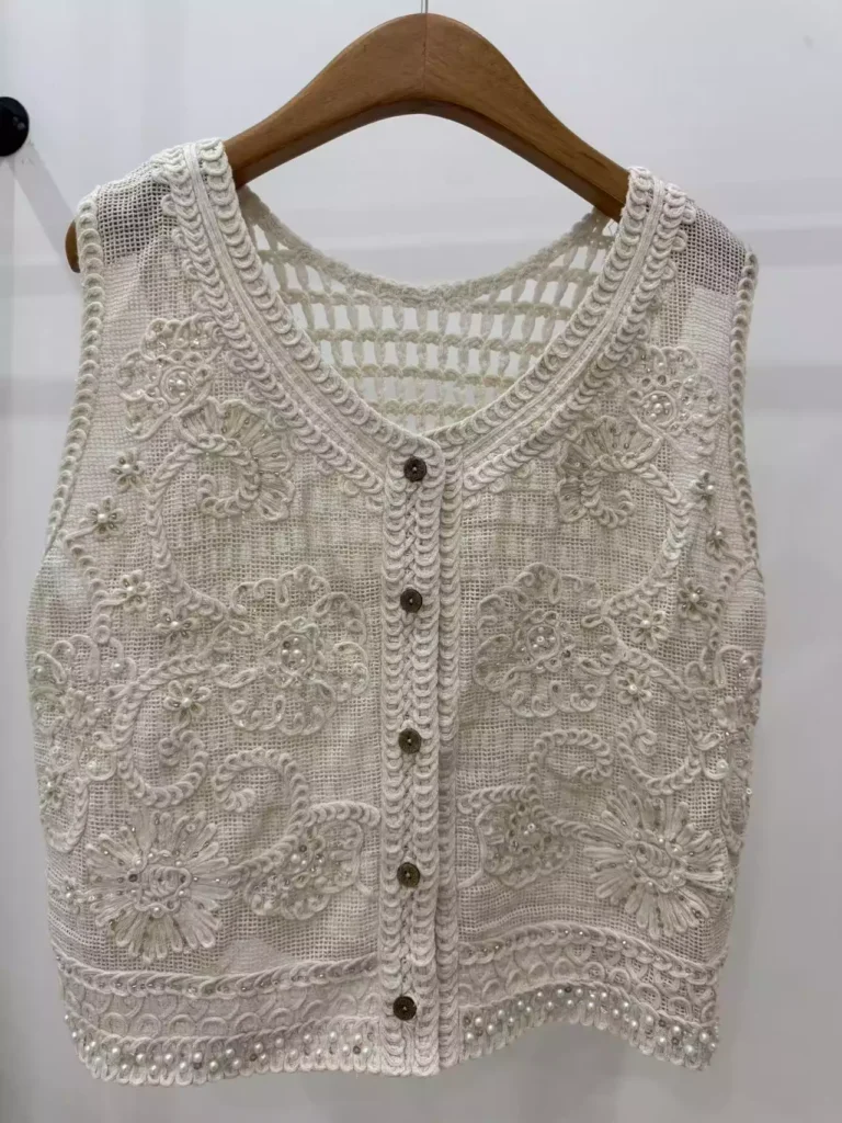 A hanger shot of a white crochet vest, showcasing intricate swirl-pattern embroidery, pearl embellishments, and a button-front design.