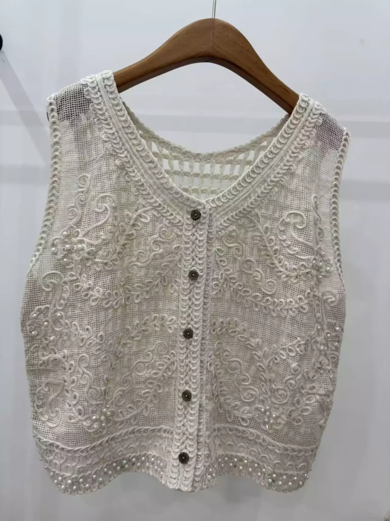 A hanger shot of an off-white crochet vest, showcasing intricate swirl-pattern embroidery, pearl embellishments, and button-front details.