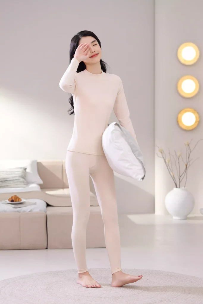 A smiling woman holding a pillow in a bright bedroom, wearing a beige mock-neck thermal underwear set, demonstrating its comfortable and casual fit for home lounging.