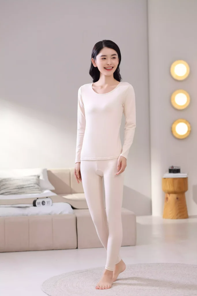 Front view of a smiling woman wearing a beige thermal underwear set, featuring a long-sleeve top and leggings with a slim-fit design in a cozy bedroom setting.