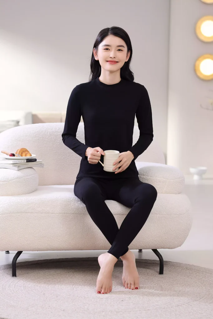 A smiling woman sitting on a white sofa in a cozy bedroom, wearing a black mock-neck thermal underwear set, demonstrating its comfortable and casual fit for home lounging.