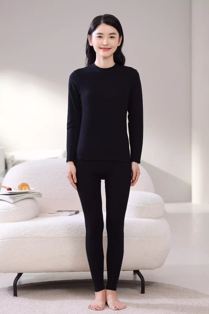Front view of a woman wearing a black mock-neck thermal underwear set, featuring a long-sleeve top and leggings with a slim-fit design in a cozy bedroom setting.