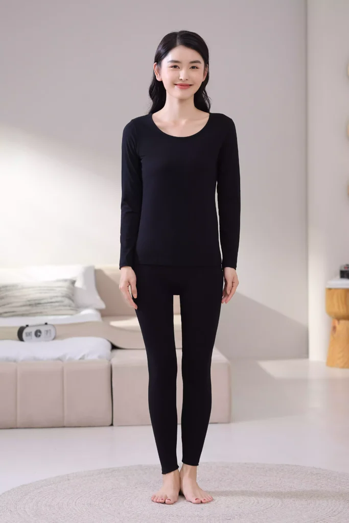 Front view of a woman wearing a black thermal underwear set, featuring a long-sleeve top and leggings with a slim-fit design in a cozy bedroom.