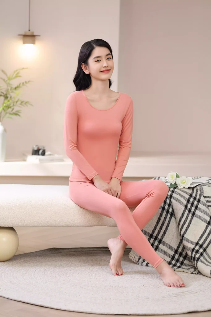 A smiling woman sitting comfortably on a bench in a cozy bedroom, wearing a coral pink thermal underwear set, highlighting its soft, casual fit for loungewear.