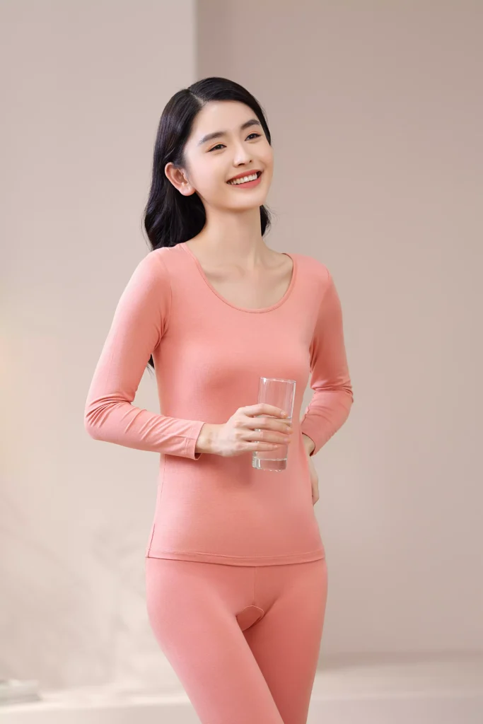 Front view of a smiling woman holding a glass of water, wearing a coral pink thermal underwear set with a slim-fit design in a cozy, bright room.