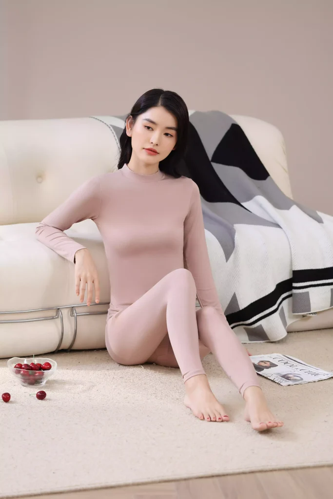 A woman sitting on the floor leaning against a white sofa, wearing a dusty pink mock-neck thermal underwear set, showcasing its casual and comfortable fit for home life.