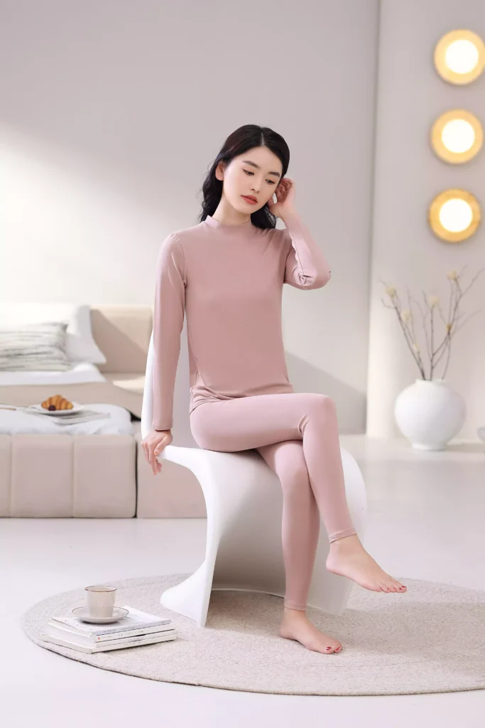 A woman sitting on a white chair in a bright, cozy bedroom, wearing a dusty pink mock-neck thermal underwear set, demonstrating its comfortable and casual fit for home lounging.