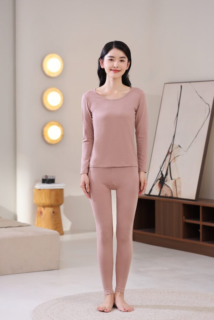 Front view of a woman wearing a dusty pink thermal underwear set, showcasing the slim-fit top and leggings in a cozy, bright bedroom setting.