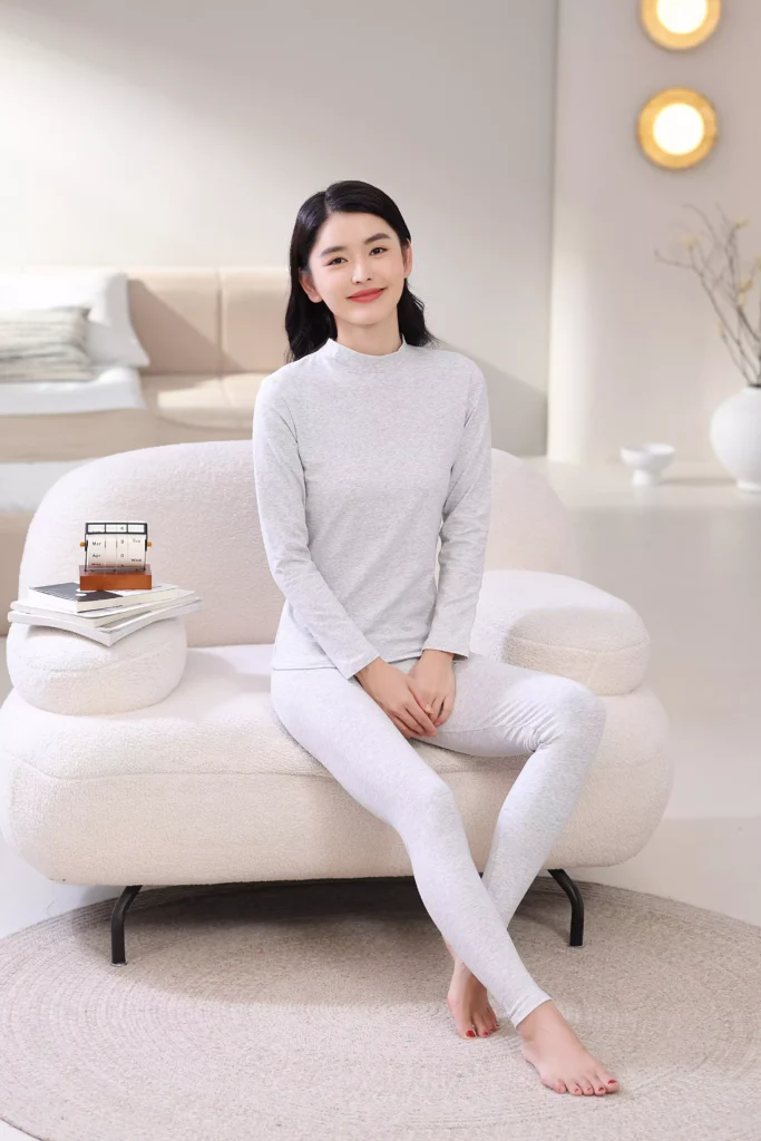 A smiling woman sitting on a white sofa in a bright bedroom, wearing a grey mock-neck thermal underwear set, demonstrating its comfortable and casual fit for home lounging