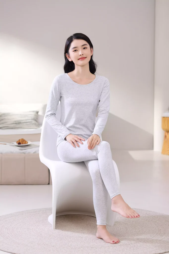 A smiling woman sitting on a white chair in a bright bedroom, wearing a grey thermal underwear set, demonstrating its comfortable and casual fit for home lounging.