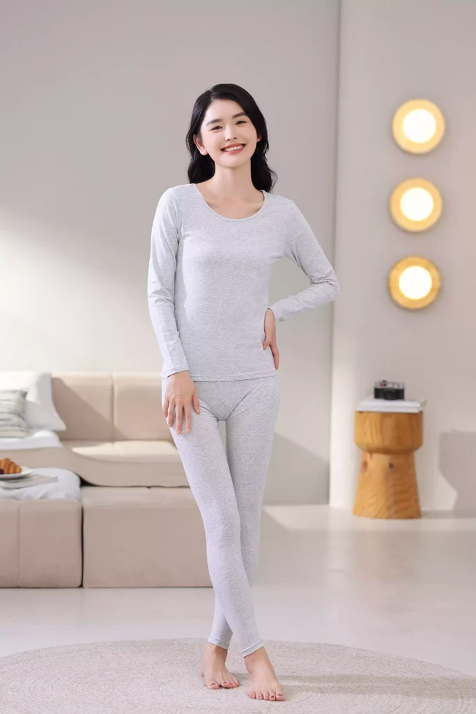Front view of a smiling woman wearing a grey thermal underwear set, featuring a long-sleeve top and leggings with a slim-fit design in a cozy bedroom setting.