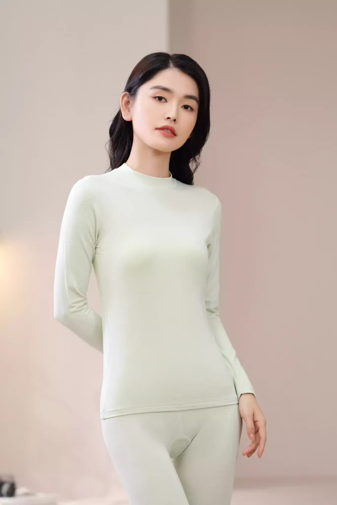 Front view of a woman wearing a light green mock-neck thermal underwear set, featuring a long-sleeve top and leggings with a slim-fit design in a cozy interior setting.
