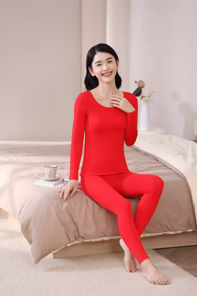 A smiling woman sitting on a bed in a bright bedroom, wearing a vibrant red thermal underwear set, demonstrating its cozy and casual fit for home lounging.