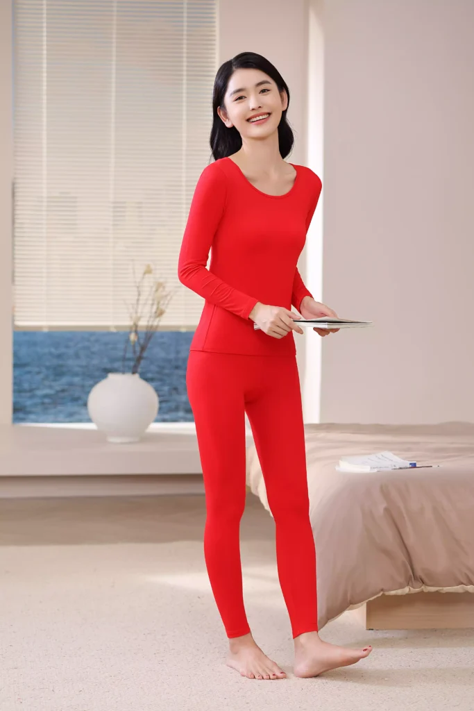 Front view of a smiling woman wearing a vibrant red thermal underwear set, featuring a long-sleeve top and leggings with a slim-fit design in a cozy bedroom setting.