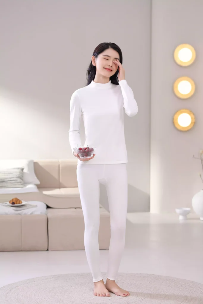 A smiling woman wearing a white mock-neck thermal underwear set, featuring a long-sleeve top and leggings in a bright, cozy bedroom setting.