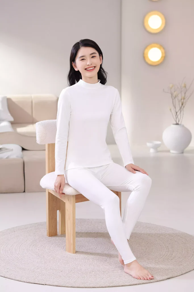 A smiling woman wearing a white mock-neck thermal underwear set, sitting on a chair in a bright bedroom, demonstrating its comfortable fit for home lounging.