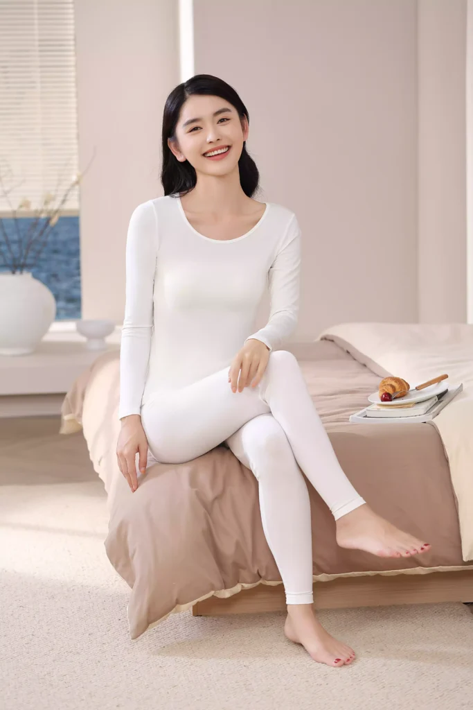 A smiling woman wearing a white thermal underwear set while sitting on a bed in a cozy, bright bedroom, showcasing the comfortable and casual fit of the loungewear.